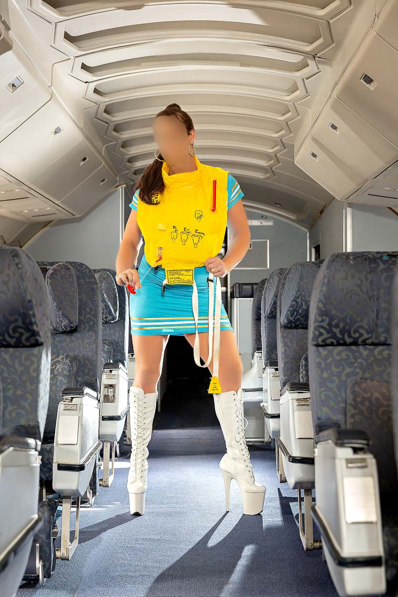Lily Levine Geelong cougar escort in the isle of a 747 doing a safety demonstration with a yellow lifejacket. Wearing a light blue skintight busty blue dress with high white boots laced and a 70's ponytail