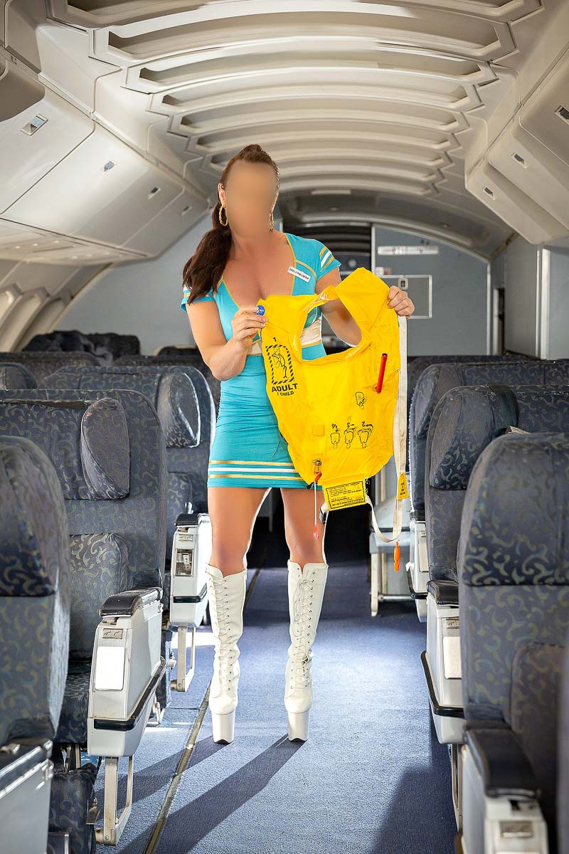 Lily Levine Geelongs best escort companion in the isle of a jumbo jet doing a safety demonstration with a yellow lifejacket. Wearing light blue skintight airhostess outfit with thigh high lace white boots and high 70's ponytail